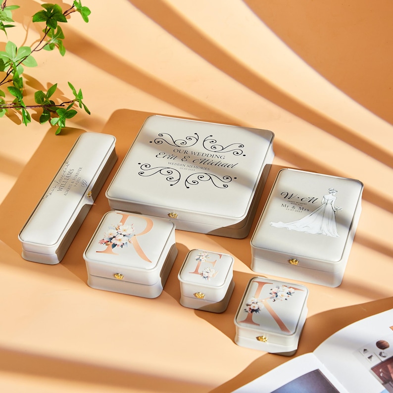 May include: A collection of ivory-colored keepsake boxes in various sizes. Some boxes feature floral designs, initials, or the text "OUR WEDDING Erin & Michael WEDDING MEMORIES". The boxes have gold-colored clasps and are arranged on a light-colored surface.