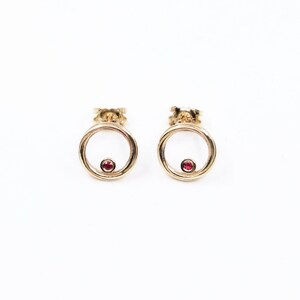 May include: A pair of gold earrings with a small red gemstone in the centre of each circle.
