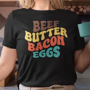 Op de afbeelding: Een zwart T-shirt met een retro-grafische afbeelding met de tekst "BEEF BUTTER BACON EGGS" in kleurrijke letters.