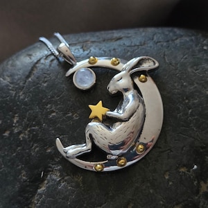 May include: A silver pendant featuring a rabbit seated on a crescent moon, holding a gold star. The moon has gold accents and a small, round, light blue stone. The pendant hangs from a silver chain.