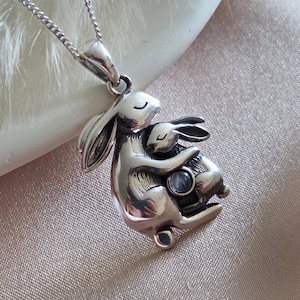 May include: A silver pendant necklace featuring a mother rabbit embracing a baby rabbit. The pendant has a detailed, three-dimensional design with a small, round gemstone. The necklace chain is delicate and silver.