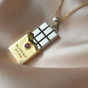 Puede incluir: Un collar con colgante de oro y plata con un dise&ntilde;o de tableta de chocolate. El colgante tiene un envoltorio dorado con el texto "AU CHOCOLAT EST. 2024" y una gema roja. La tableta de chocolate es plateada con cuadrados negros.