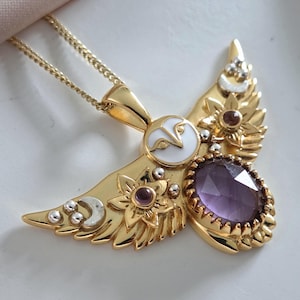 May include: Gold-coloured pendant necklace with an owl design, white enamel, and a large, oval purple gemstone. The pendant has floral and silver-coloured details. The necklace has a fine chain, suitable for everyday wear.