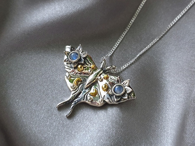 May include: A silver-toned moth pendant necklace with a delicate chain. The pendant features intricate details, including gold accents, blue gemstone accents, and floral designs. The moth's wings are spread, and the necklace rests on a gray fabric.