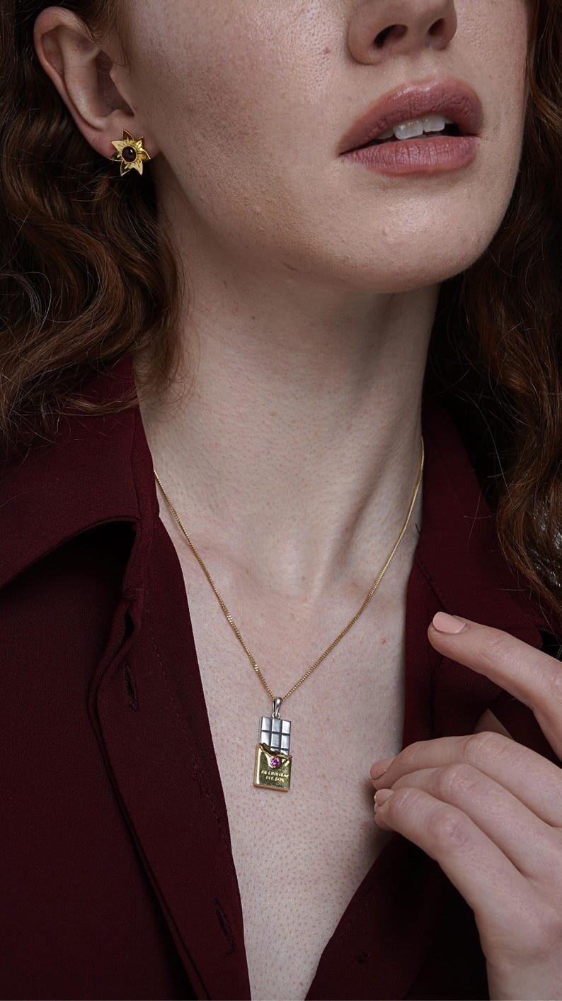 Puede incluir: Un collar dorado con un colgante en forma de tableta de chocolate, con la parte superior plateada y la inferior dorada con texto. Se ve un pendiente dorado en forma de estrella a juego con una gema roja. La persona lleva una camisa granate.