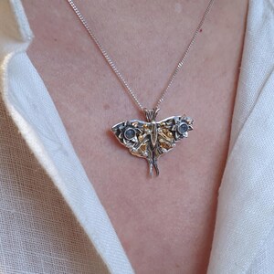 May include: A silver pendant necklace featuring a detailed moth design with gold accents and two blue gemstones. The pendant hangs from a delicate silver chain. The necklace is worn against a white linen shirt.