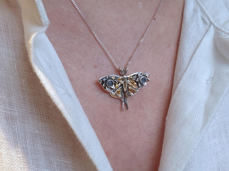 May include: A silver pendant necklace featuring a detailed moth design with gold accents and two blue gemstones. The pendant hangs from a delicate silver chain. The necklace is worn against a white linen shirt.