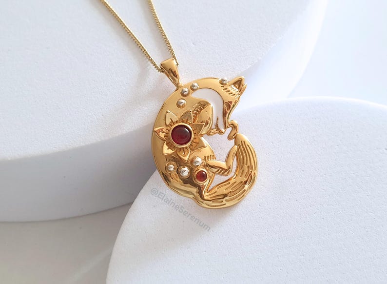 May include: Gold necklace with a pendant featuring a curled fox with red gemstones and white enamel accents. The pendant is detailed with a floral design.