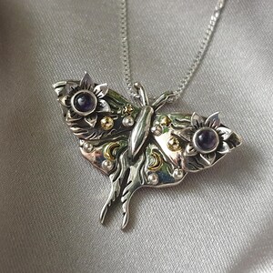 May include: A silver-toned pendant necklace featuring a detailed butterfly design. The butterfly has floral accents with purple gemstone centers and gold-colored embellishments. The necklace is displayed on a silver satin background.