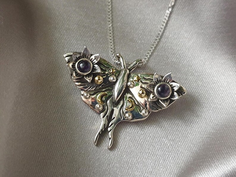 May include: A silver-toned pendant necklace featuring a detailed butterfly design. The butterfly has floral accents with purple gemstone centers and gold-colored embellishments. The necklace is displayed on a silver satin background.