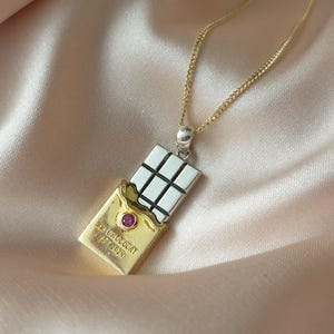 Puede incluir: Un collar con colgante de oro y plata con un dise&ntilde;o de tableta de chocolate. El colgante tiene una tableta de chocolate plateada con un envoltorio dorado, una peque&ntilde;a gema roja y el texto "Au Chocolat 1937-2024". El collar est&aacute; en una cadena de oro.
