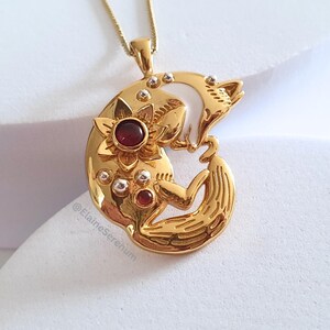 May include: Gold pendant necklace featuring a stylized fox curled around a crescent moon with a red gemstone flower. The pendant is detailed with small white beads.