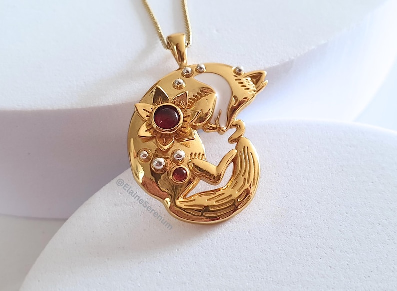 May include: Gold pendant necklace featuring a stylized fox curled around a crescent moon with a red gemstone flower. The pendant is detailed with small white beads.