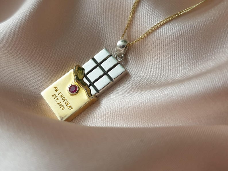 Puede incluir: Un collar con colgante de oro y plata con un dise&ntilde;o de tableta de chocolate. El colgante tiene un envoltorio dorado con el texto "AU CHOCOLAT EST. 2024" y una gema roja. La tableta de chocolate es plateada con cuadrados negros.