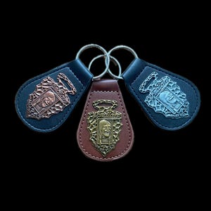 以下が含まれることがあります： 金属製のエンブレムが付いた3つの革製キーホルダー。キーホルダーは、銅、銀、金色のトーンで「Indian Motorcycles」のロゴを特徴としています。各キーホルダーには、キーを取り付けるための金属リングが付いています。キーホルダーは黒い背景に設定されています。