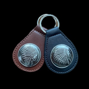 May include: Two leather keychains, one brown and one navy blue, each featuring a silver-colored coin with a Native American chief profile. The keychains have a teardrop shape and a metal ring for attaching keys.