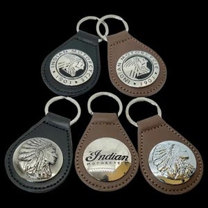 Könnte beinhalten: Fünf Leder-Schlüsselanhänger mit silberfarbenen Metall-Emblemen. Die Schlüsselanhänger sind aus schwarzem und braunem Leder gefertigt und zeigen das Indian Motorcycle-Logo oder ein indianisches Profil. Jeder Schlüsselanhänger hat einen Metallring zum Befestigen von Schlüsseln.