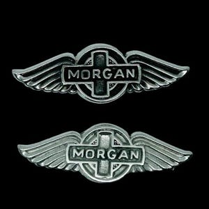 May include: Two silver metal badges with a winged design. Each badge has a cross shape with the name "MORGAN" inscribed. The wings are detailed with feather patterns, set against a black backdrop.