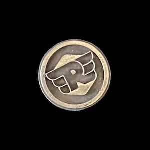 May include: A circular, silver-coloured metal button with a raised design. The design features a stylized letter "R" with wings inside a circular border. The button is set against a black background.