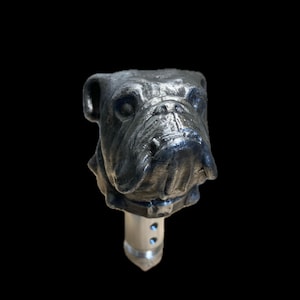 May include: A silver bulldog head bottle stopper with a silver metal base. The bulldog has a realistic design with a furrowed brow and a slightly open mouth.