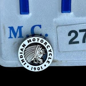 Peut inclure: Gros plan sur un emblème argenté Indian Motorcycle avec une image en noir et blanc d'une coiffe amérindienne. L'emblème est fixé à une plaque d'immatriculation blanche avec le texte "M.C. 27".