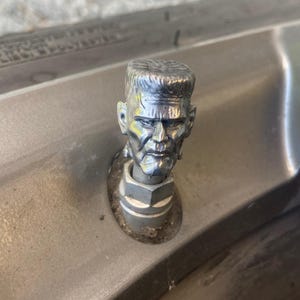 May include: A silver metal Frankenstein head figurine. The figurine is attached to a metal base with a bolt. The head has detailed features, including a furrowed brow and prominent cheekbones. The figurine is mounted on a grey surface.