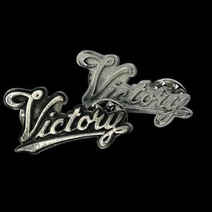Puede incluir: Dos pines de metal plateados con la palabra "Victory" en relieve en cada uno. Los pines están ligeramente superpuestos.