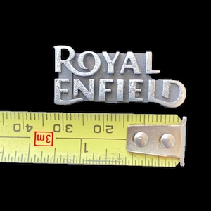 Pode incluir: Um emblema de metal prateado com as palavras "ROYAL ENFIELD" em letras maiúsculas. O emblema tem aproximadamente 7,6 cm de comprimento. Uma fita métrica amarela está colocada por baixo.