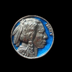 May include: A silver-colored coin with a Native American profile and the year 1913. The coin has a blue enamel border and the word "LIBERTY" is visible. The coin is set against a black background.