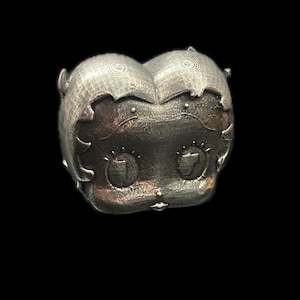 Könnte beinhalten: Eine silberfarbene Metallsculptur eines Comic-Charakters. Die Skulptur ist mit einer strukturierten Oberfläche detailliert und zeigt ein stilisiertes Gesicht mit großen Augen und einem kleinen Mund.