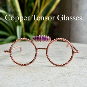 Puede incluir: Copper Tensor Glasses con monturas circulares hechas de alambre de cobre trenzado. Un pequeño cristal cilíndrico de color púrpura está envuelto en alambre de cobre y colocado en el centro del puente. El texto Copper Tensor Glasses es visible.