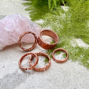 May include: A collection of four copper rings featuring various designs, including two twisted bands, one wide smooth band, and one thin plain band. The rings are arranged near a large, rough-cut piece of pale pink rose quartz crystal.