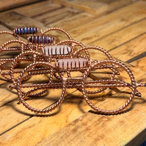 May include: A collection of four decorative wire-wrapped spectacles crafted from twisted copper wire. Each pair features a unique cylindrical gemstone set horizontally across the bridge, including blue, purple, white, and pale pink stones.
