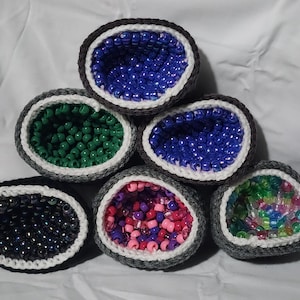 May include: Six crocheted geode-shaped items are displayed. Each has a grey exterior with a white border, filled with colourful beads. Bead colours include blue, green, black, pink, purple, and a mix of colours. The geodes are handmade.