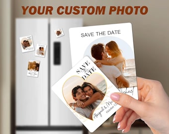 May include: Two white save the date magnets with gold accents. The magnets feature a couple in a heart-shaped frame and a couple embracing on a beach. The text on the magnets reads "Save the Date" and "Abigail & Michael, October 12".