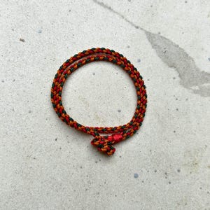 May include: A braided rope bracelet in a circular shape. The bracelet is made of red, green, and gold threads. The bracelet is knotted at the end. The bracelet is on a light gray surface.