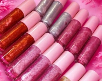 May include: An assortment of lip gloss tubes in various shades of pink, silver, and bronze. The tubes have pink caps and are arranged on a pink surface. The lip glosses appear to contain glitter and shimmer.