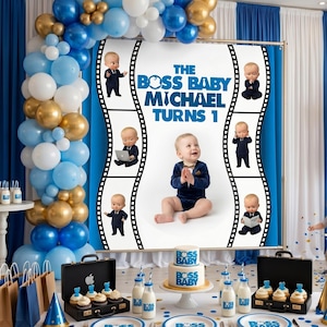 Puede incluir: Una escena de fiesta de cumpleaños con globos azules, dorados y blancos, una pancarta que dice "The Boss Baby Michael Turns 1" y una tarta. La tarta tiene escrito "Boss Baby". Hay cupcakes, botellas de leche y un maletín.