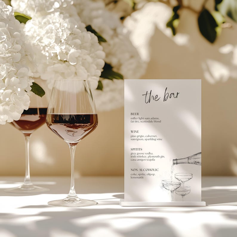 May include: Two wine glasses filled with red wine stand before a white floral arrangement. A small sign reads "the bar" and lists drink options including beer, wine, spirits, and non-alcoholic beverages.