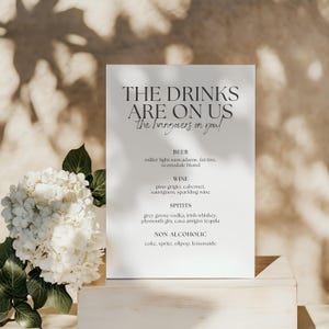 May include: A white rectangular sign with the text "THE DRINKS ARE ON US" and a list of drink options. The sign is propped up on a wooden surface, with white flowers and greenery in the foreground.