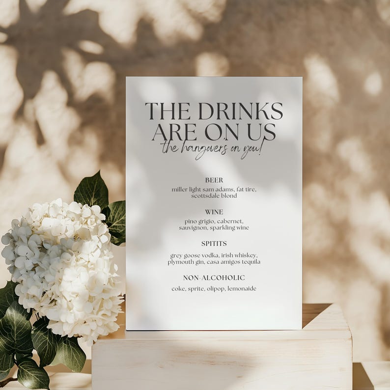 May include: A white rectangular sign with the text "THE DRINKS ARE ON US" and a list of drink options. The sign is propped up on a wooden surface, with white flowers and greenery in the foreground.