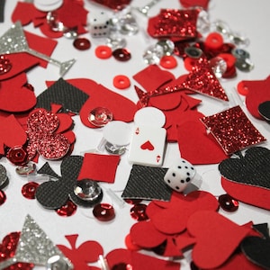 May include: A collection of red, black, and silver casino-themed confetti, including hearts, spades, clubs, diamonds, dice, and playing cards. Some pieces have glitter accents. Small, red, and silver sequins are scattered throughout.