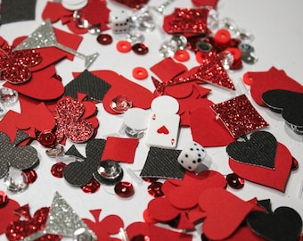 May include: A collection of red, black, and silver casino-themed confetti, including hearts, spades, clubs, diamonds, dice, and playing cards. Some pieces have glitter accents. Small, red, and silver sequins are scattered throughout.