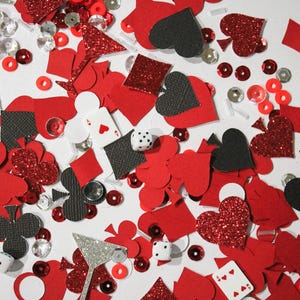 May include: A scattered assortment of red, black, and silver decorative elements. The image includes playing card suits, dice, sequins, and glitter shapes, creating a festive and playful theme. The items are scattered on a white surface.