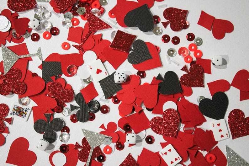 May include: A scattered assortment of red, black, and silver decorative elements. The image includes playing card suits, dice, sequins, and glitter shapes, creating a festive and playful theme. The items are scattered on a white surface.