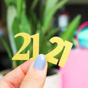 May include: A gold glittery cutout of the number 21. The surface of the number has a textured, sparkling finish that reflects light. The cutout is shaped as a single piece featuring the digits two and one, designed for decorative purposes.