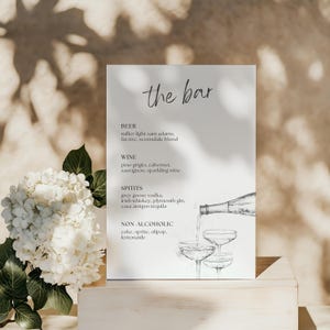 May include: A white bar menu sign with the words "the bar" in script. The menu lists beer, wine, spirits, and non-alcoholic drinks. A sketched illustration shows a bottle pouring into three glasses. A white flower arrangement is in the background.