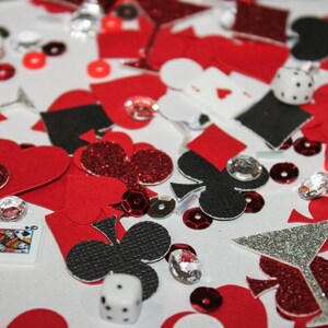 May include: A scattered assortment of red, black, and silver glitter confetti, including hearts, clubs, diamonds, and spades. Dice, playing cards, and sequins are also present, creating a casino or party theme.