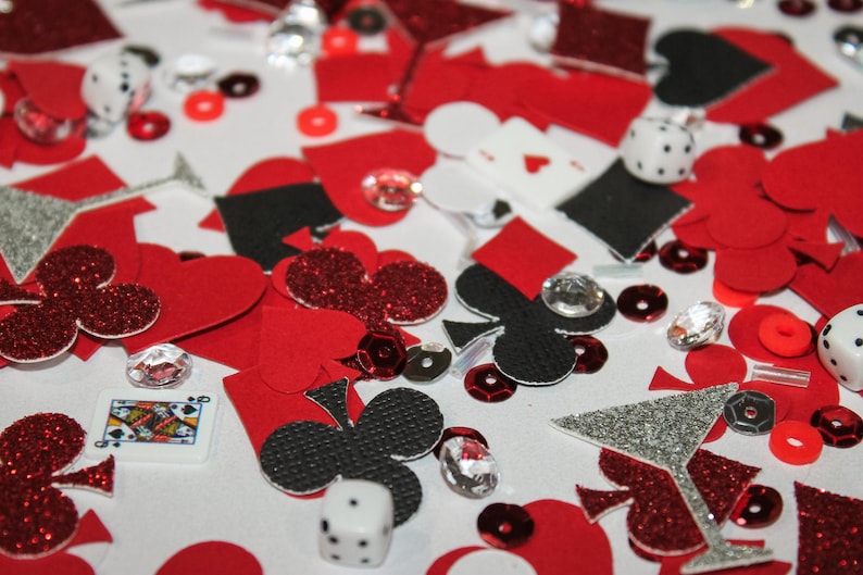 May include: A scattered assortment of red, black, and silver glitter confetti, including hearts, clubs, diamonds, and spades. Dice, playing cards, and sequins are also present, creating a casino or party theme.