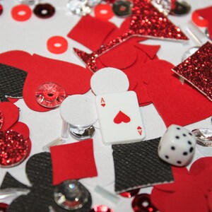 May include: A collection of red, black, and white casino-themed decorations. Includes playing card suit shapes, a die, an Ace of Hearts playing card, and sparkling embellishments. The items are scattered on a white surface.